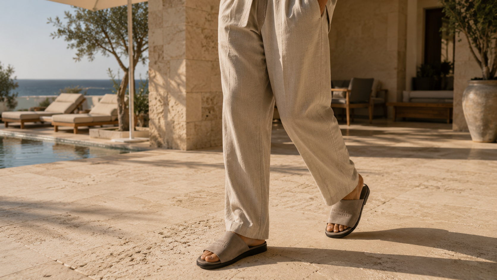 Persona con pantalones de lino y slides de gamuza caminando junto a una piscina en un entorno mediterráneo